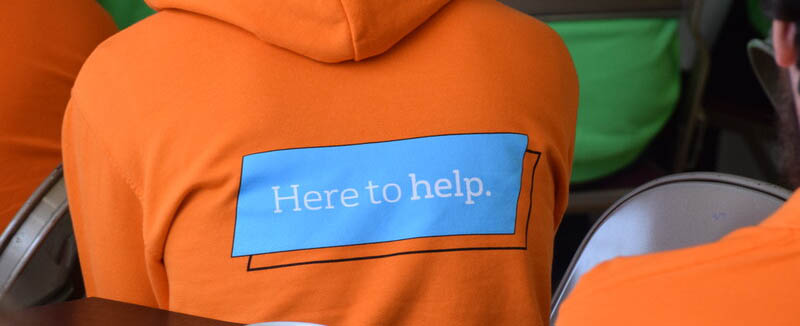 The back of a person wearing an orange hoodie with a 'here to help' slogan on it.